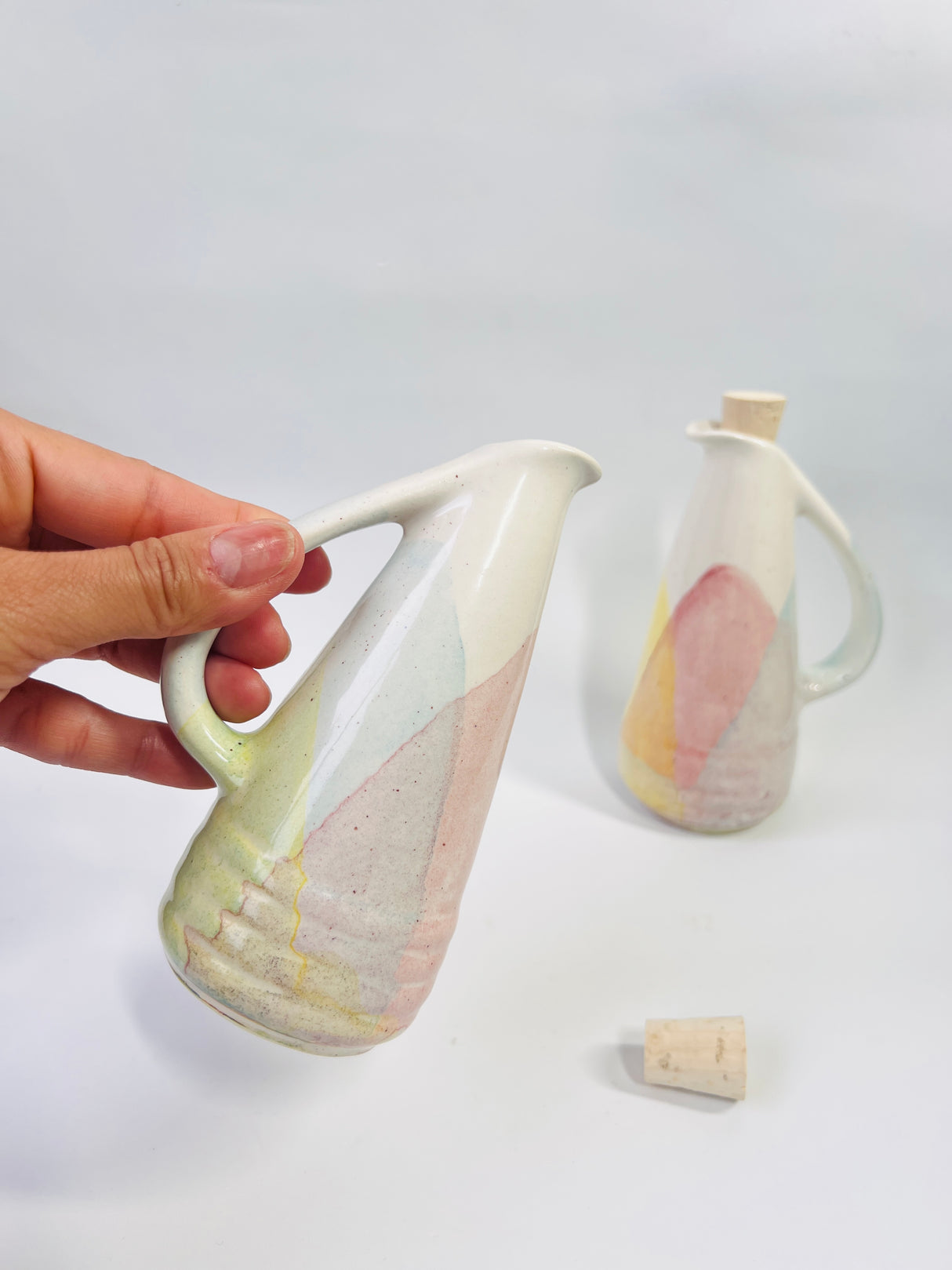 Hand holding a ceramic pitcher with a second pitcher in the background on a white surface