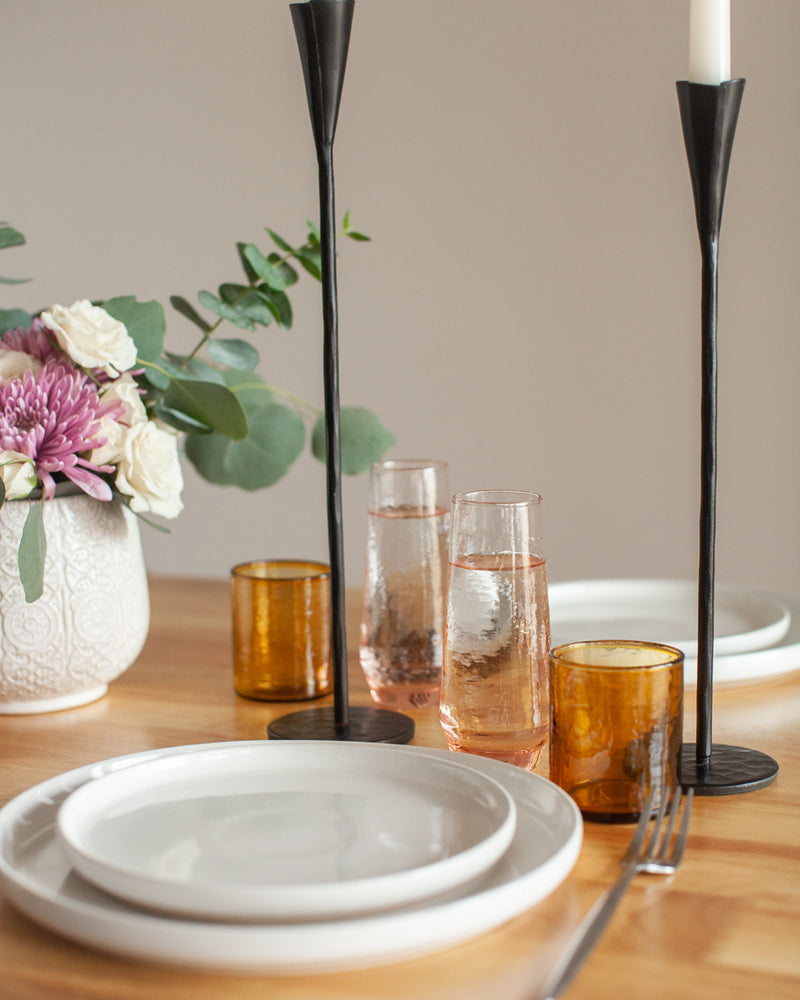 Dining table setting with white plates, amber glasses, black candle holders, and a vase with flowers.