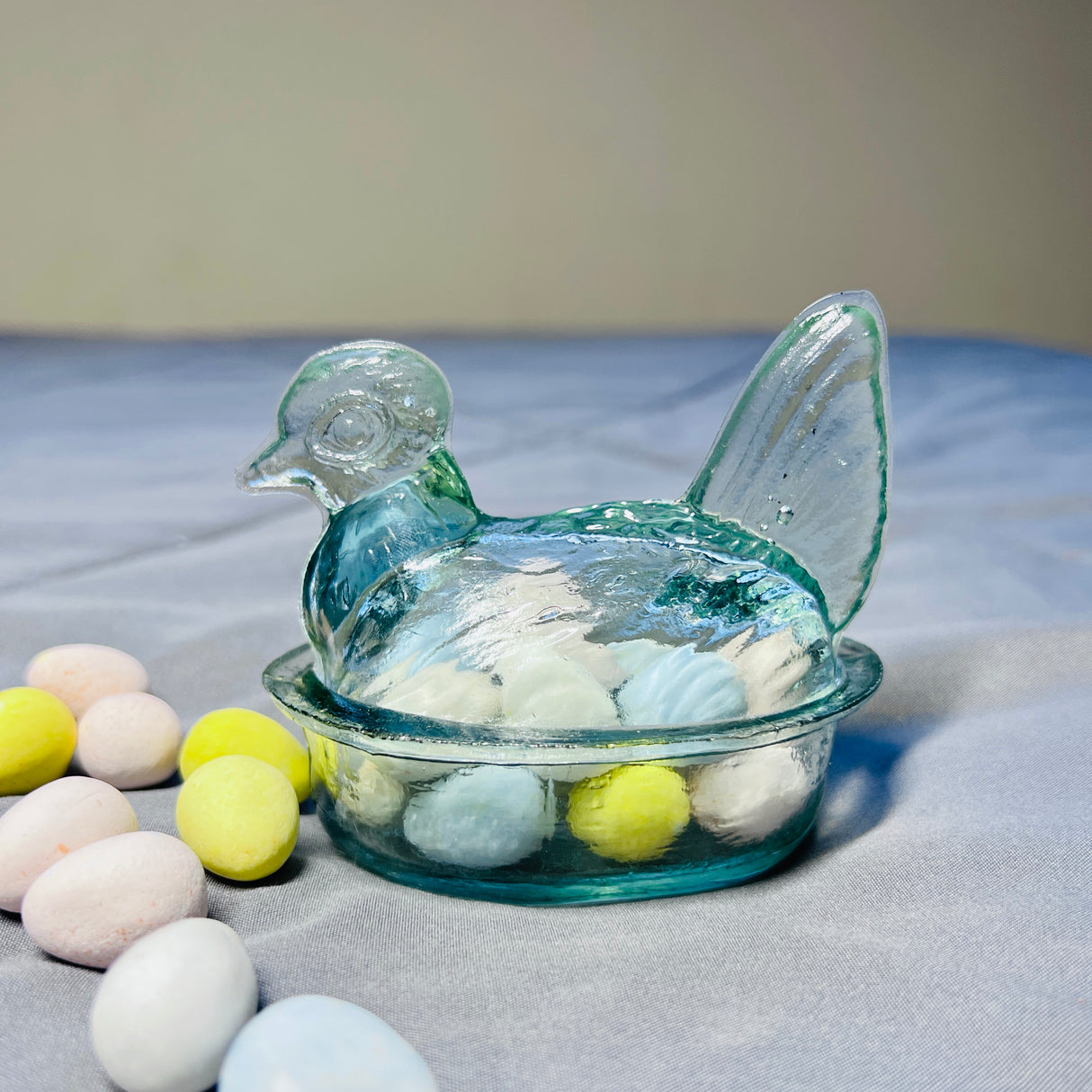 Hand blown clear glass bowl in the shape of a hen with a lid, containing smaller eggs of various colors, displayed on a grey surface.
