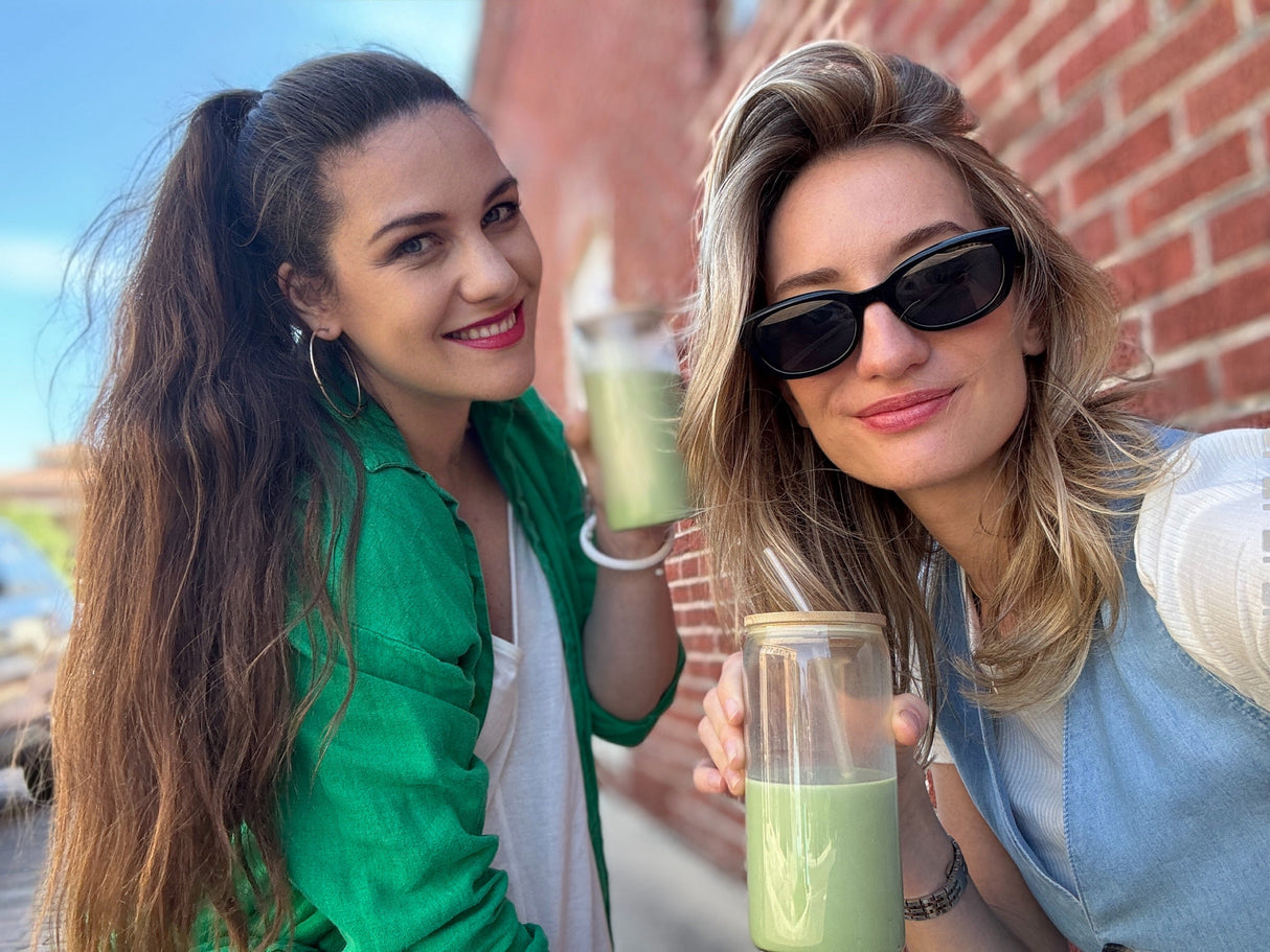 Two women smile outdoors, holding clear cups of matcha lattes made with Ceremonial Grade Matcha Latte Powder Mix with Natural Vanilla Flavor, standing in front of a brick wall.