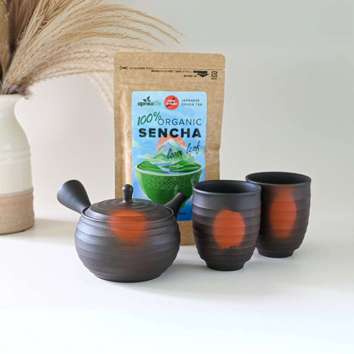 A Japanese Kyusu Set, including a 270ml teapot with filters and two teacups, rests on a pristine white surface alongside organic sencha tea, with a delicate vase adding elegance to the background.