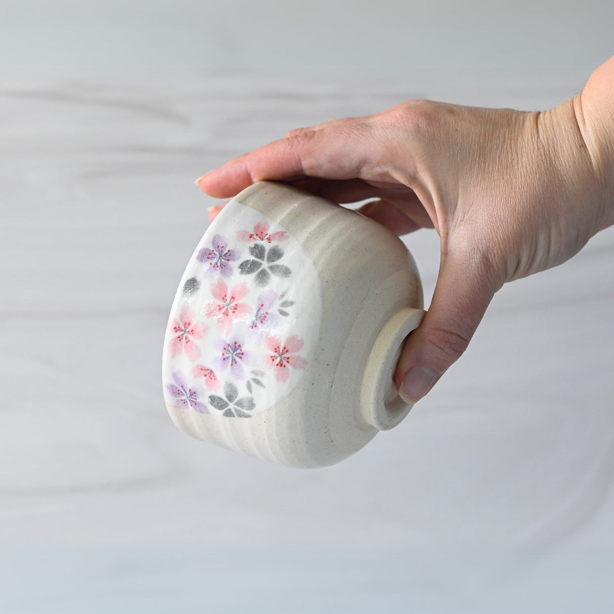 A hand cradles a Kohiki Pink Sakura Ceramic Matcha Bowl, adorned with a pink and purple floral design, reminiscent of those used in a traditional matcha tea ceremony.
