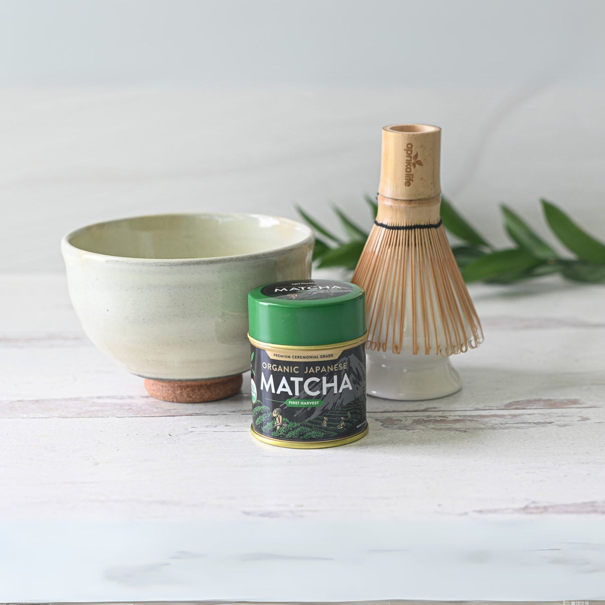The Kohiki White Clay Matcha Bowl, along with a bamboo whisk, sits on a light wooden surface with a green matcha tin nearby, framed by lush greenery.