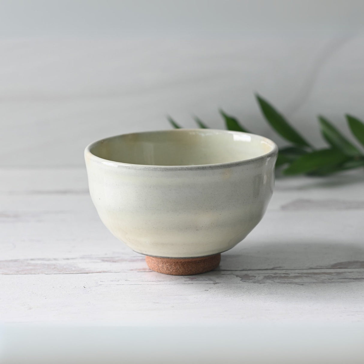 The Kohiki White Clay Matcha Bowl, with a cork base on a light wooden surface and surrounded by green leaves, highlights the elegance of Japanese ceramic craftsmanship.