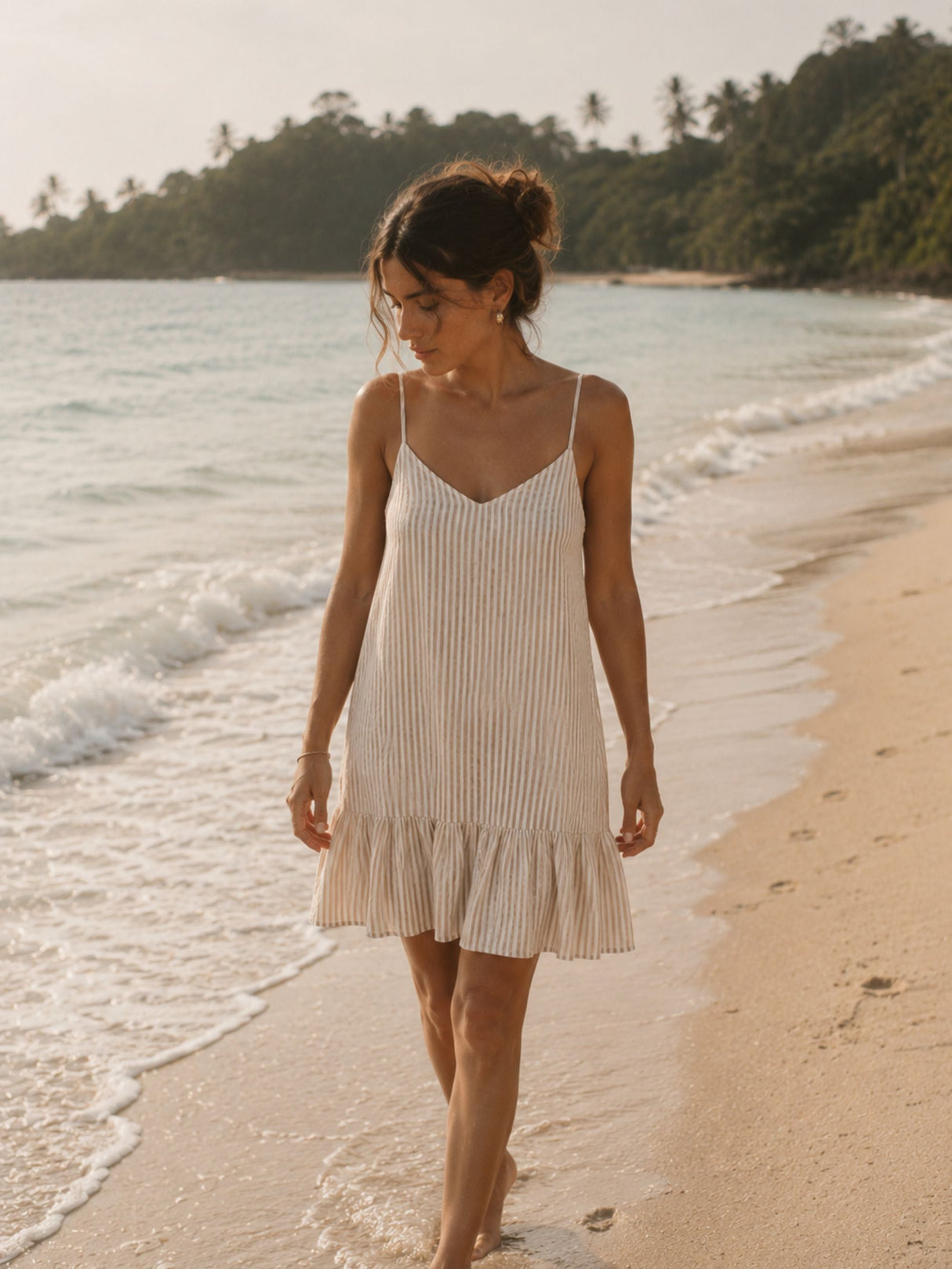 Woman in a striped dress walking on a beach with ocean waves and greenery in the background