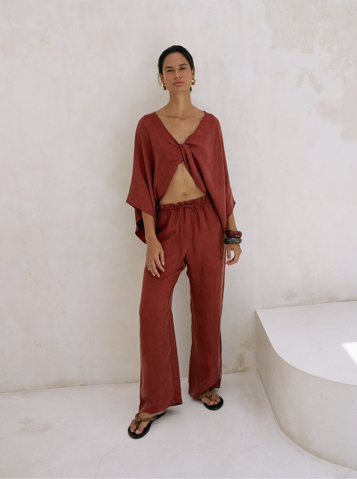 Woman wearing a rust-colored outfit standing against a white wall.