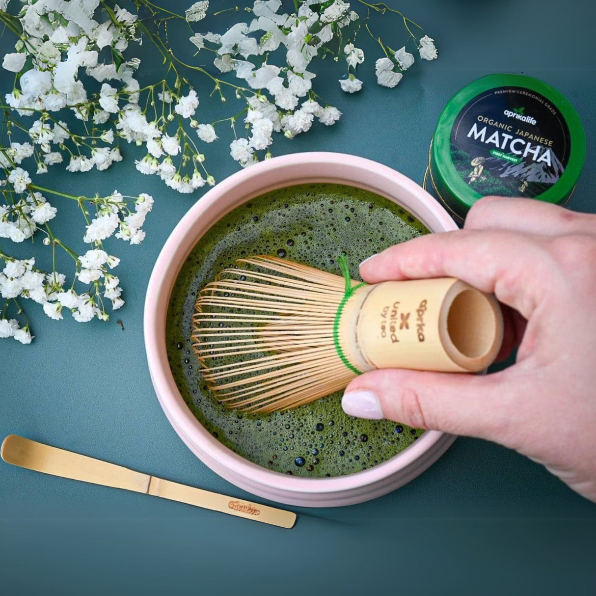 Using the Matcha Bamboo Whisk & Holder Set, enjoy hand whisking matcha in a bowl, surrounded by flowers, with a bamboo scoop nearby and a matcha container elegantly within reach.