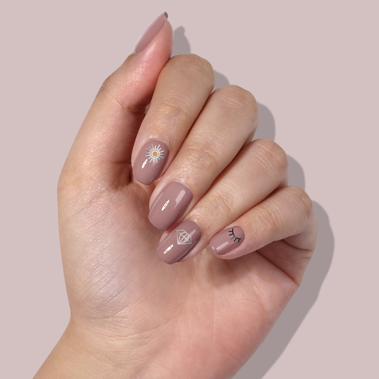 A hand with manicured nails painted in a nude shade features small sun, diamond, and eyelash designs using LONDONTOWN's Nail Decals - Cosmic, showcasing easy nail art at its finest.