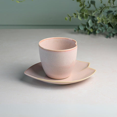 A Pink LOVE Ceramic Tea Cup with Saucer sits elegantly, accented by a leafy branch in the backdrop, evoking a serene and timeless Japanese Ippuku-wan tea moment.