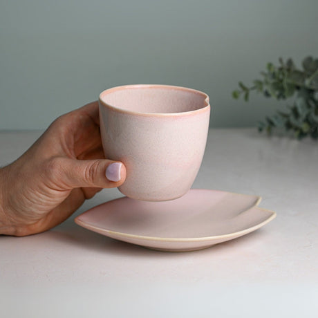 A hand holds a Pink LOVE Ceramic Tea Cup with Saucer on a light surface, against a backdrop of lush greenery.