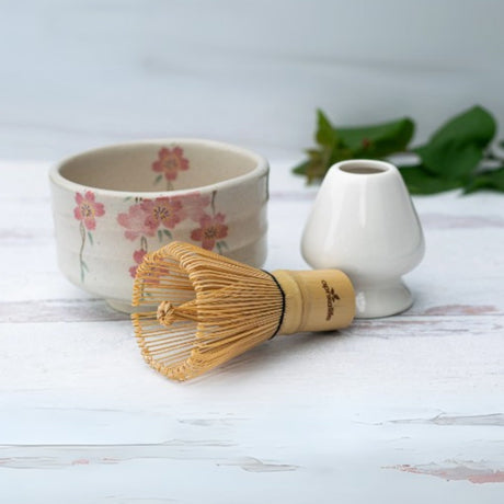 A Pink Sakura Flowers Ceramic Matcha Bowl with a Bamboo Matcha Whisk and holder rests on a white surface, accompanied by a vase and leaves. This exquisite set showcases the artistry of Japanese pottery.