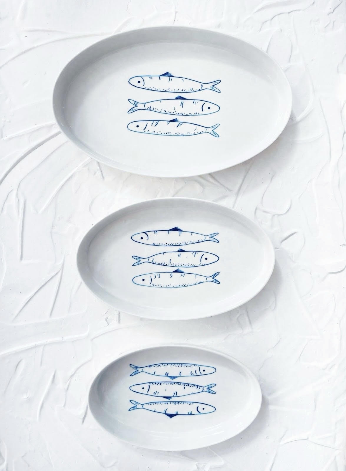 Three white plates with blue fish illustrations on a textured white background. Sardines Nesting Plates