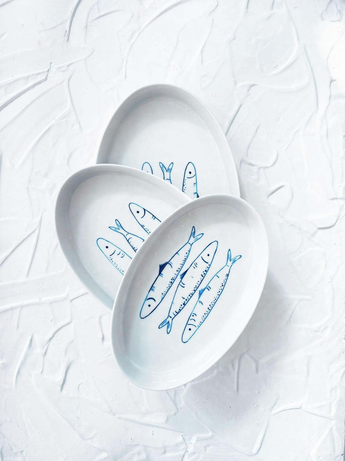 Three white plates with blue fish illustrations on a textured white background. Sardines Nesting Plates
