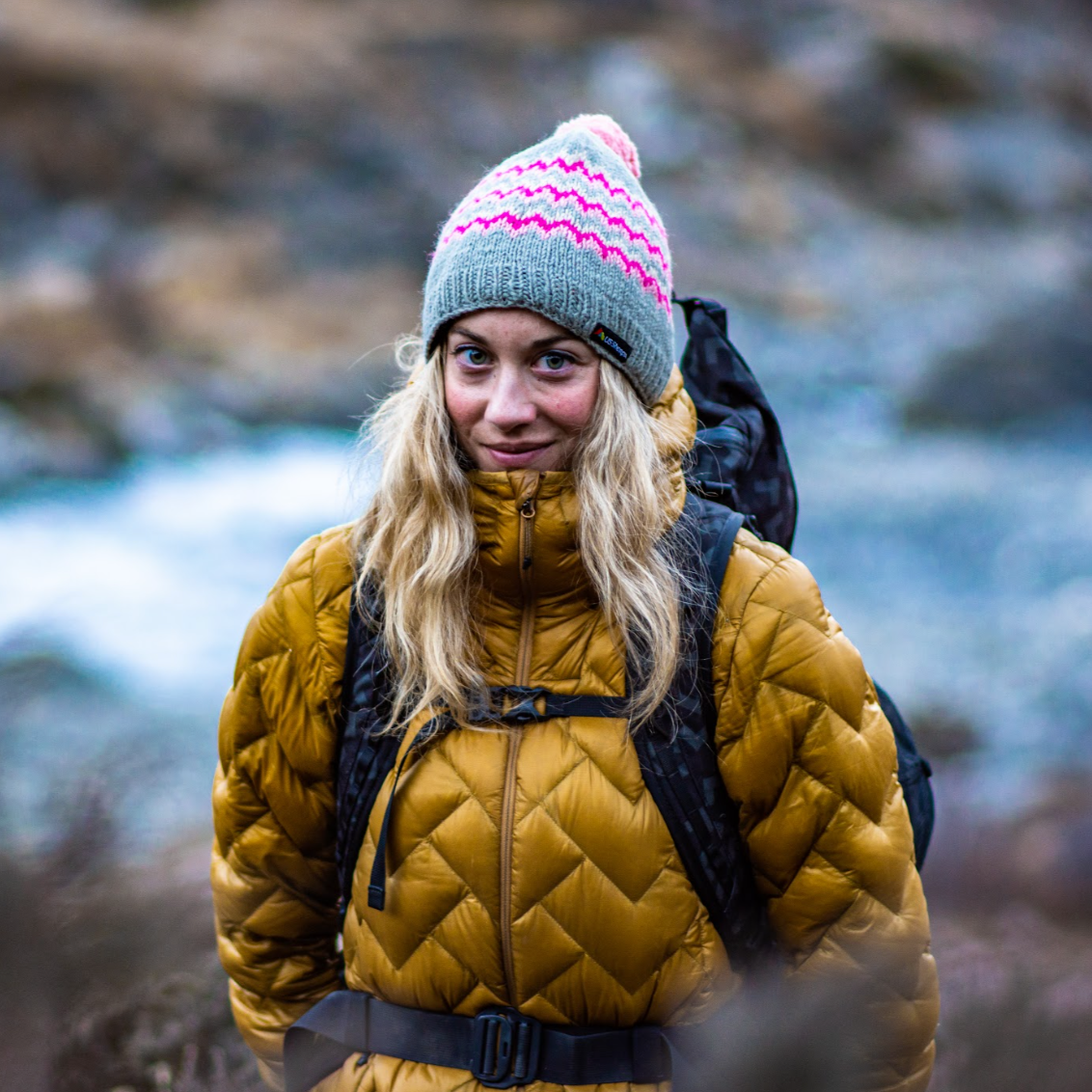 Person wearing a yellow puffer jacket and colorful knit hat standing outdoors with a blurred natural background