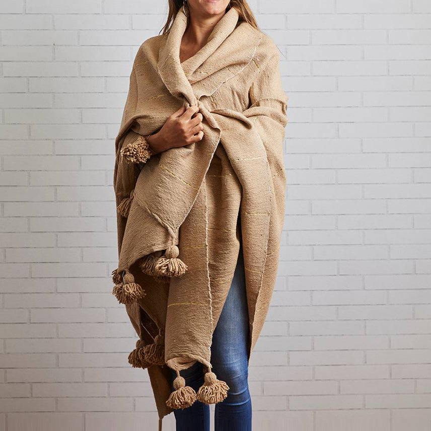 Person wearing a beige blanket wrapped around them against a white brick wall.