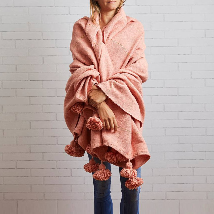 Person wearing a pink poncho with pom-poms against a light gray brick wall.