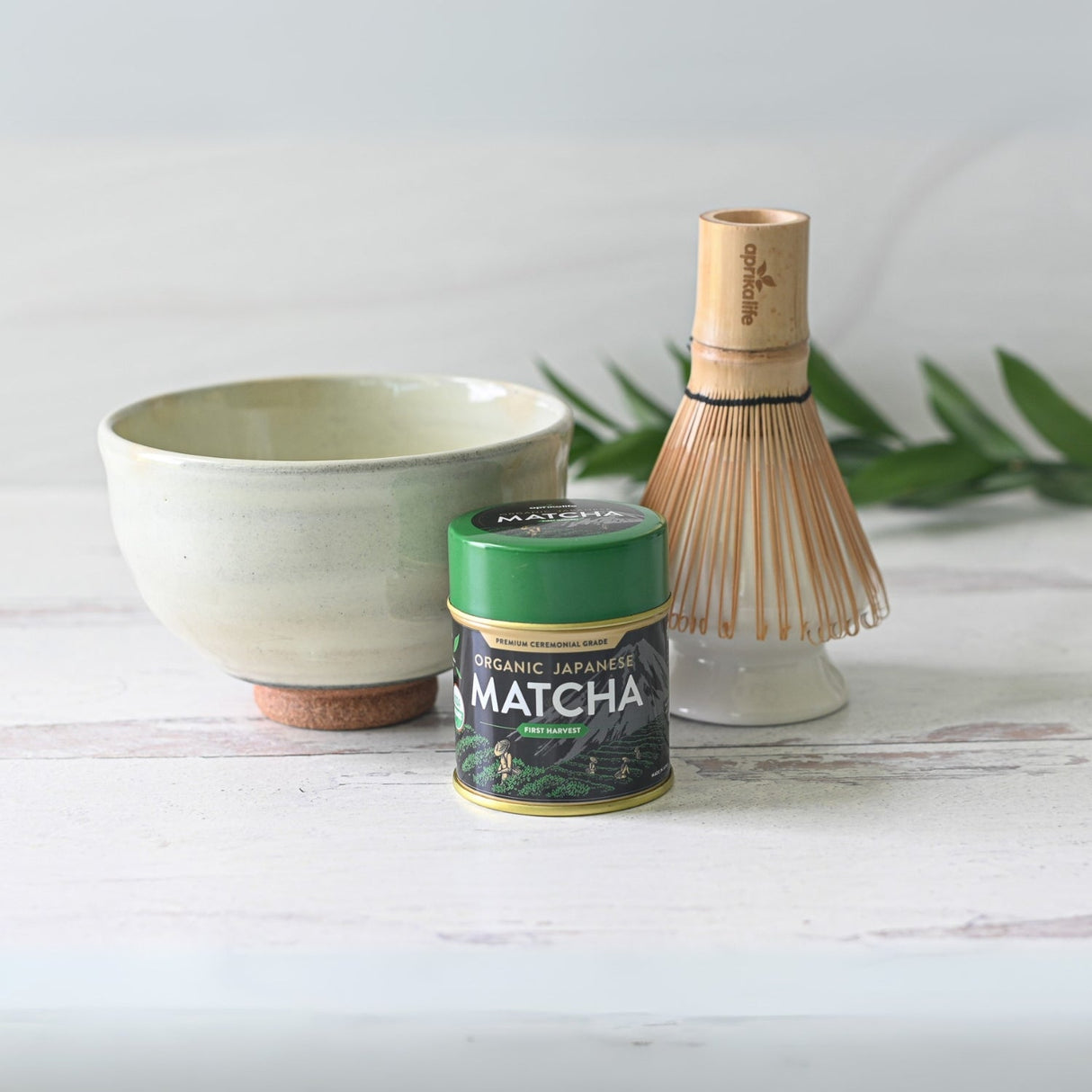White Kohiki Ceramic Matcha Bowl Set with matcha tin and bamboo whisk on a light wooden surface, surrounded by leaves.