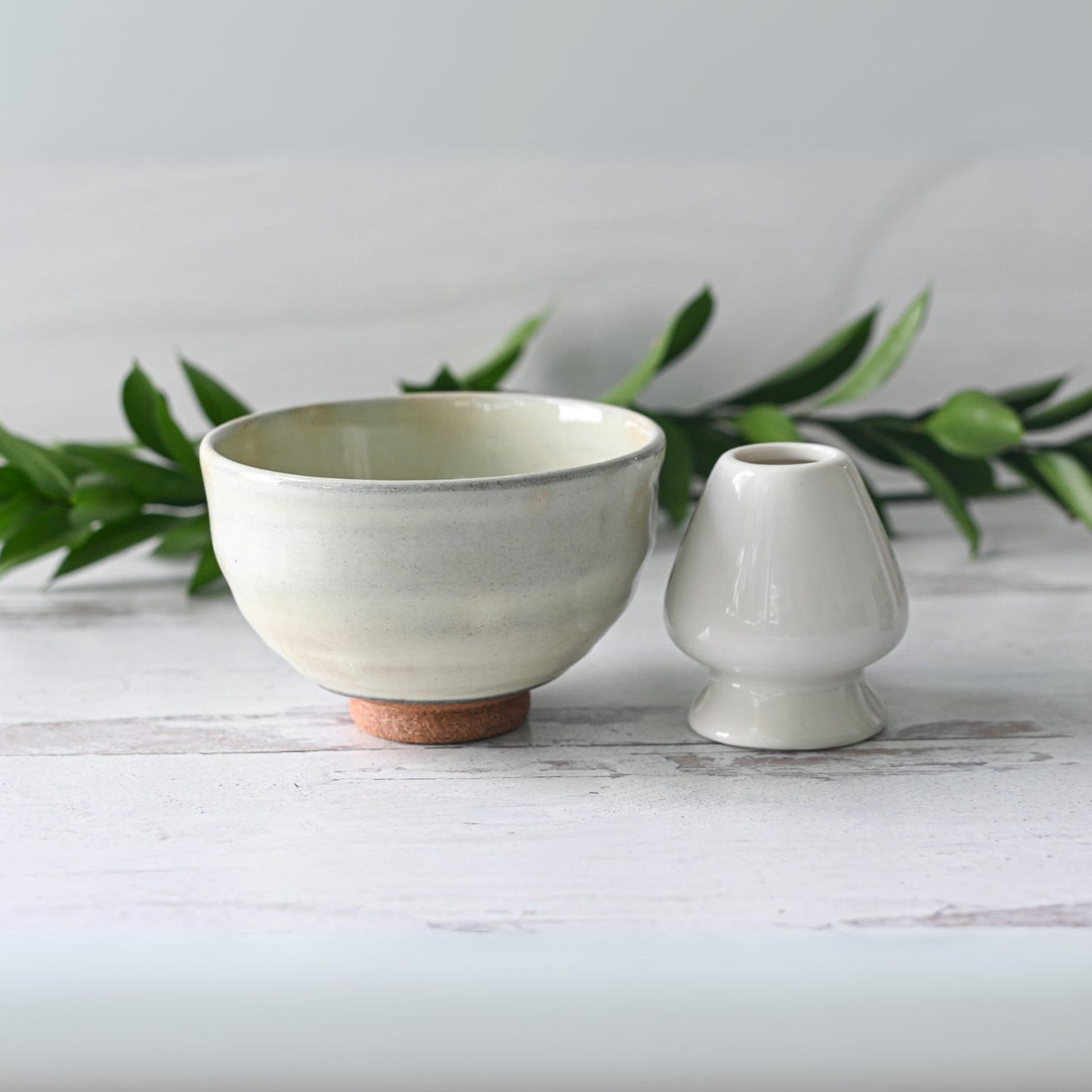 The White Kohiki Ceramic Matcha Bowl rests on a light wooden surface, accompanied by a small vase crafted with Minoyaki techniques, while green foliage sets a serene background.