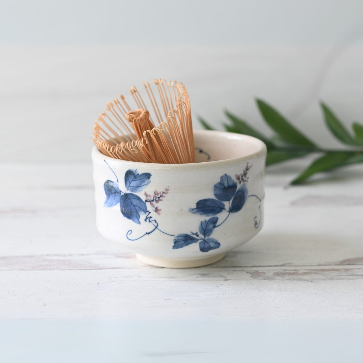 A beautiful White Clay Kudzu Flowers Matcha Bowl, complete with a bamboo whisk and holder, sits elegantly on a white wooden surface adorned with green leaves. This Organic Japanese Matcha Set is the ideal starter set for matcha enthusiasts.