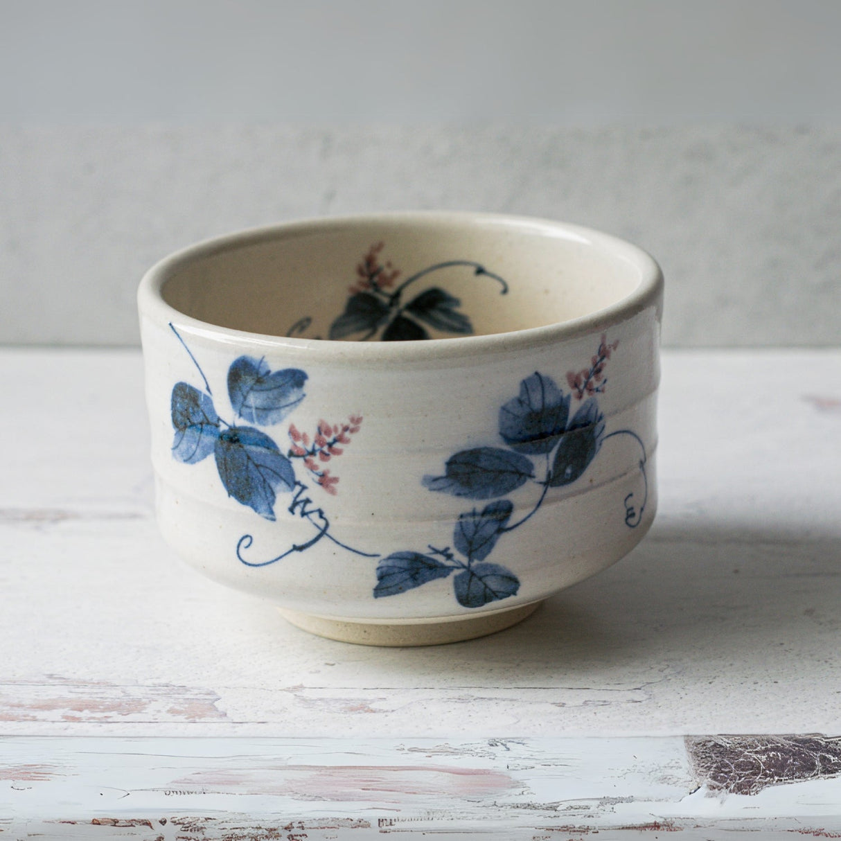 A Minoyaki pottery teacup with a blue floral vine design elegantly sits on a light wooden surface, ideal for your White Clay Kudzu Flowers Matcha Bowl Set.
