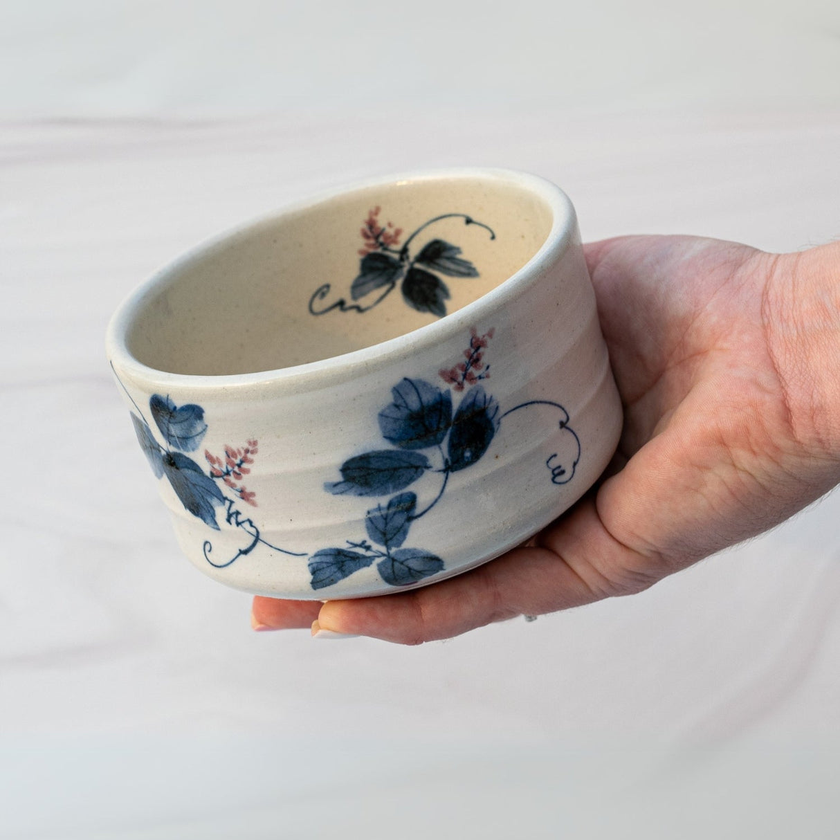 A hand delicately holds the White Clay Kudzu Flowers Matcha Bowl, featuring a blue floral design reminiscent of traditional Minoyaki pottery, set against a light background.