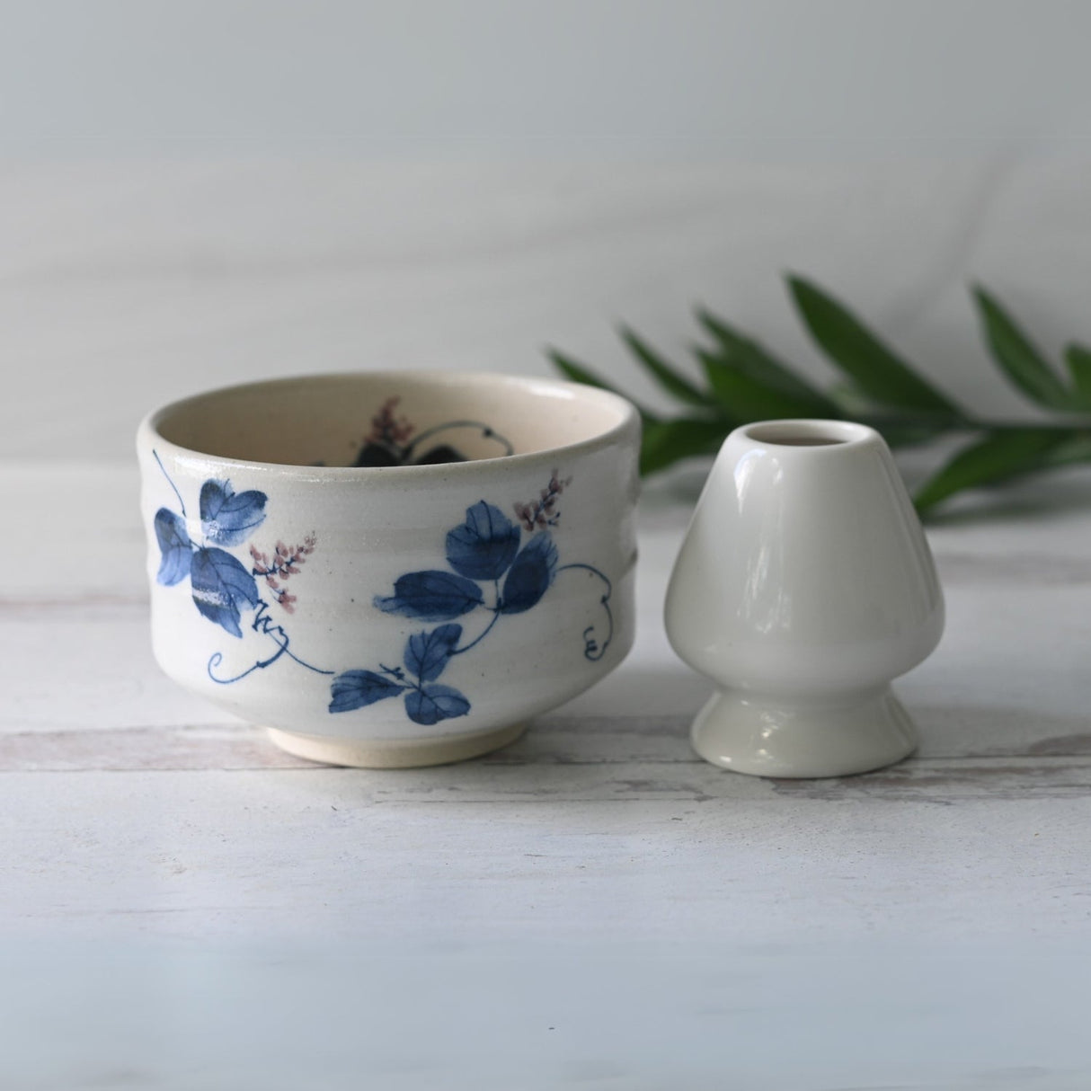 A White Clay Kudzu Flowers Matcha Bowl, part of the Bamboo Matcha Whisk, Holder, and Organic Japanese Matcha Set, sits beside a small white vase on a light wooden surface.