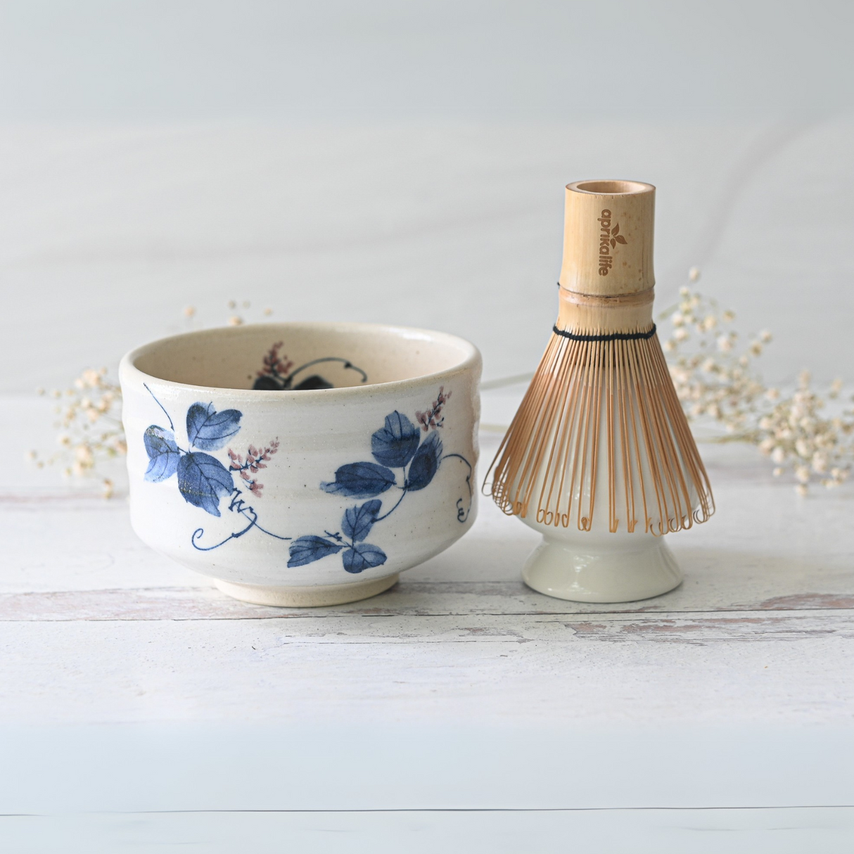 A White Clay Kudzu Flowers Matcha Bowl from the White Clay Kudzu Flowers Matcha Set sits beside a bamboo matcha whisk on a light marble surface.