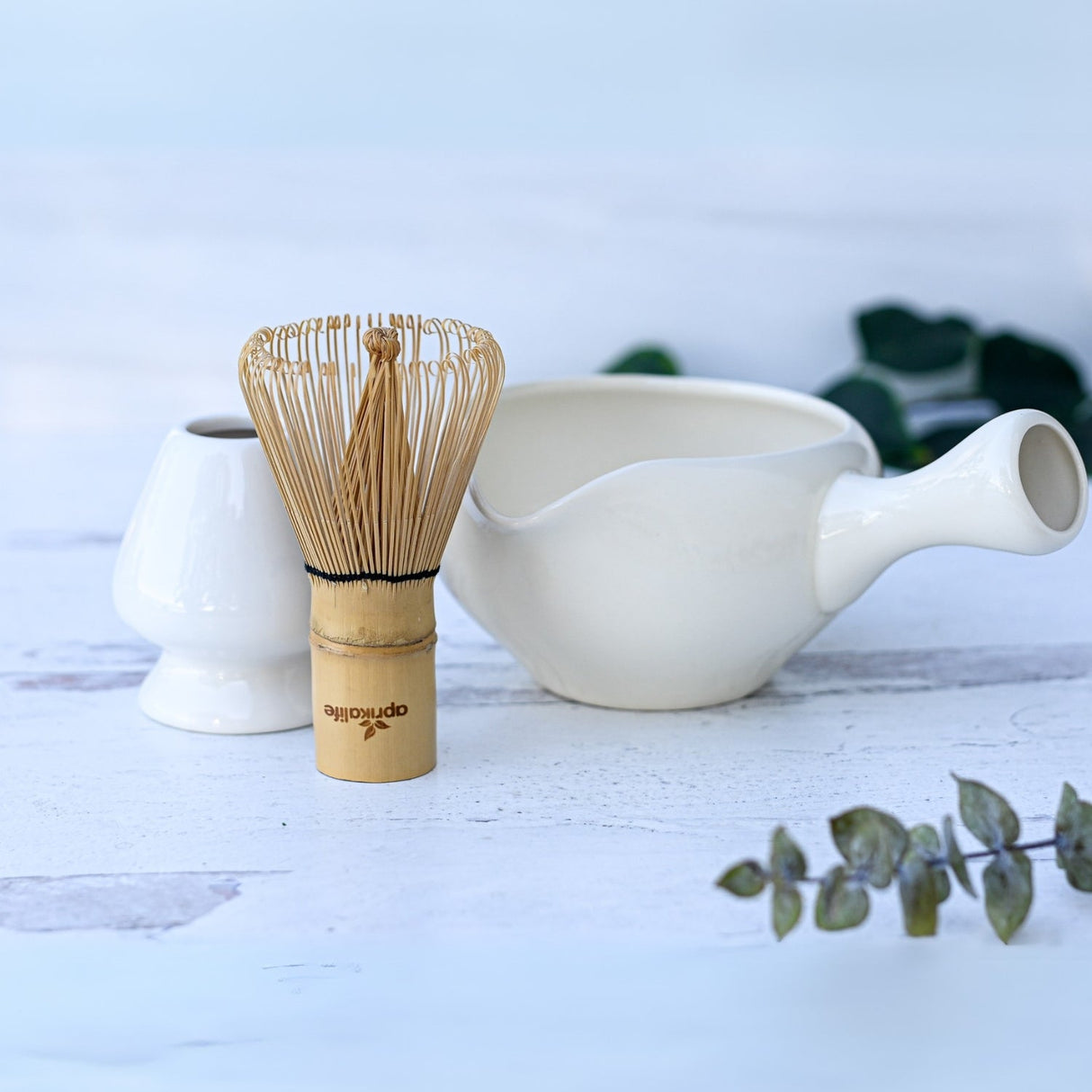 A bamboo matcha whisk sits elegantly beside a White Ceramic Matcha Bowl with Spout, framed by lush greenery. Perfect for a Japanese tea ceremony or as a thoughtful housewarming gift.