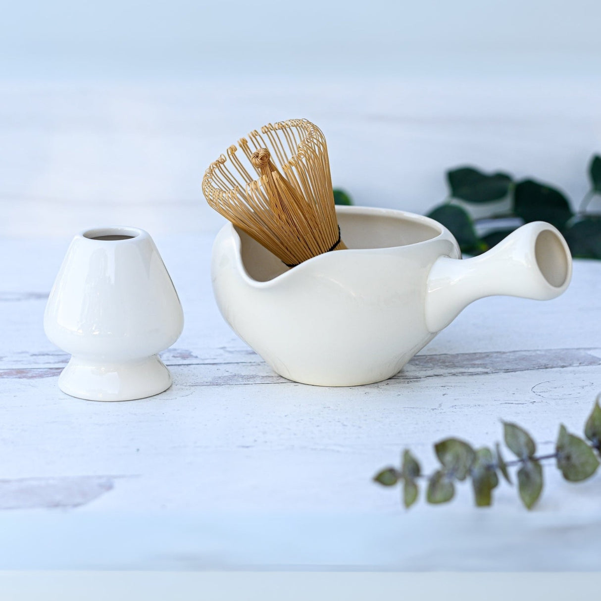 The White Ceramic Matcha Bowl with Spout, accompanied by a bamboo whisk and a small white vase on a wooden surface, makes an ideal housewarming gift.