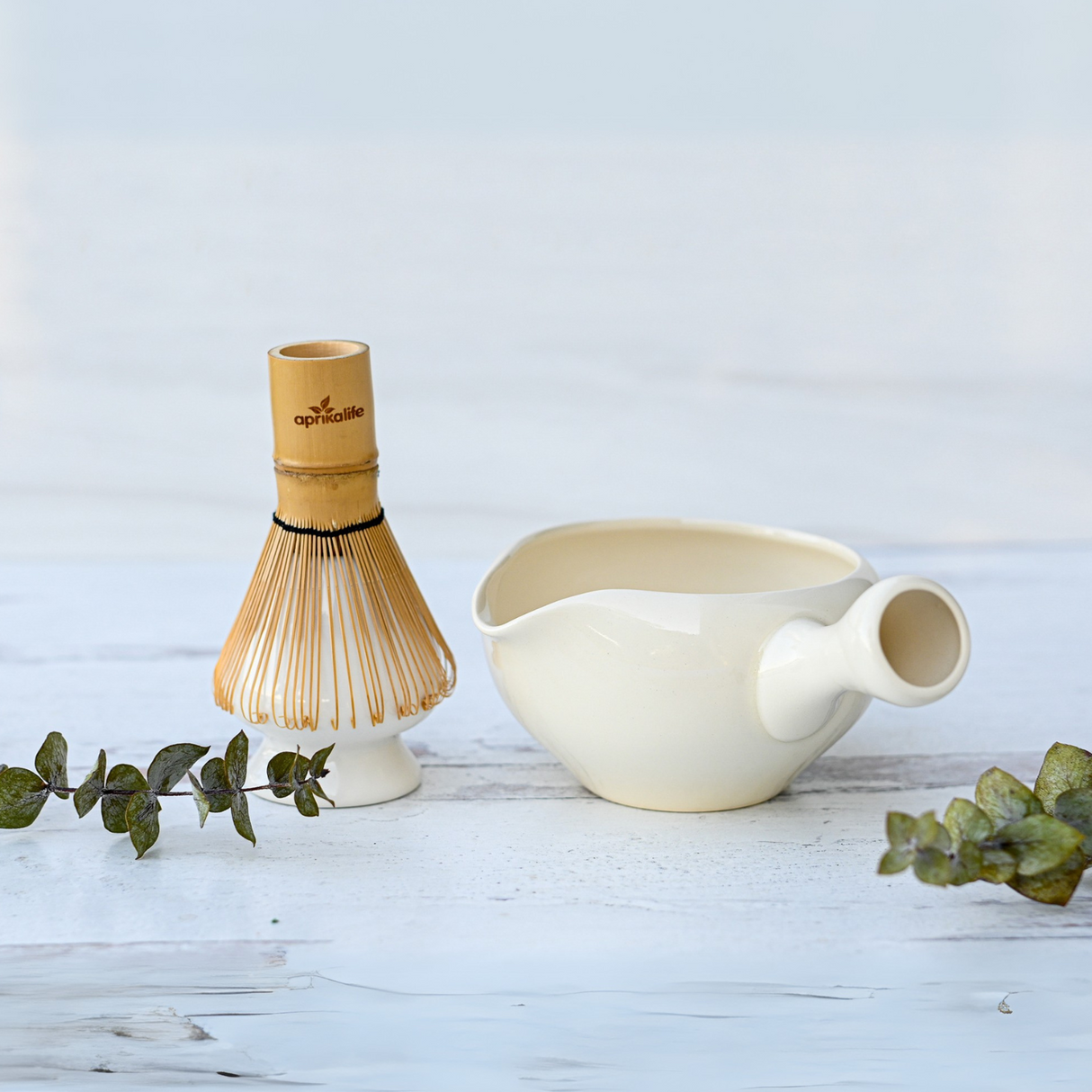 The White Ceramic Matcha Bowl with Spout, Bamboo Matcha Whisk, Whisk Holder and Organic Japanese Matcha Set rests on a white surface with green leaves—ideal for preparing authentic Japanese matcha.