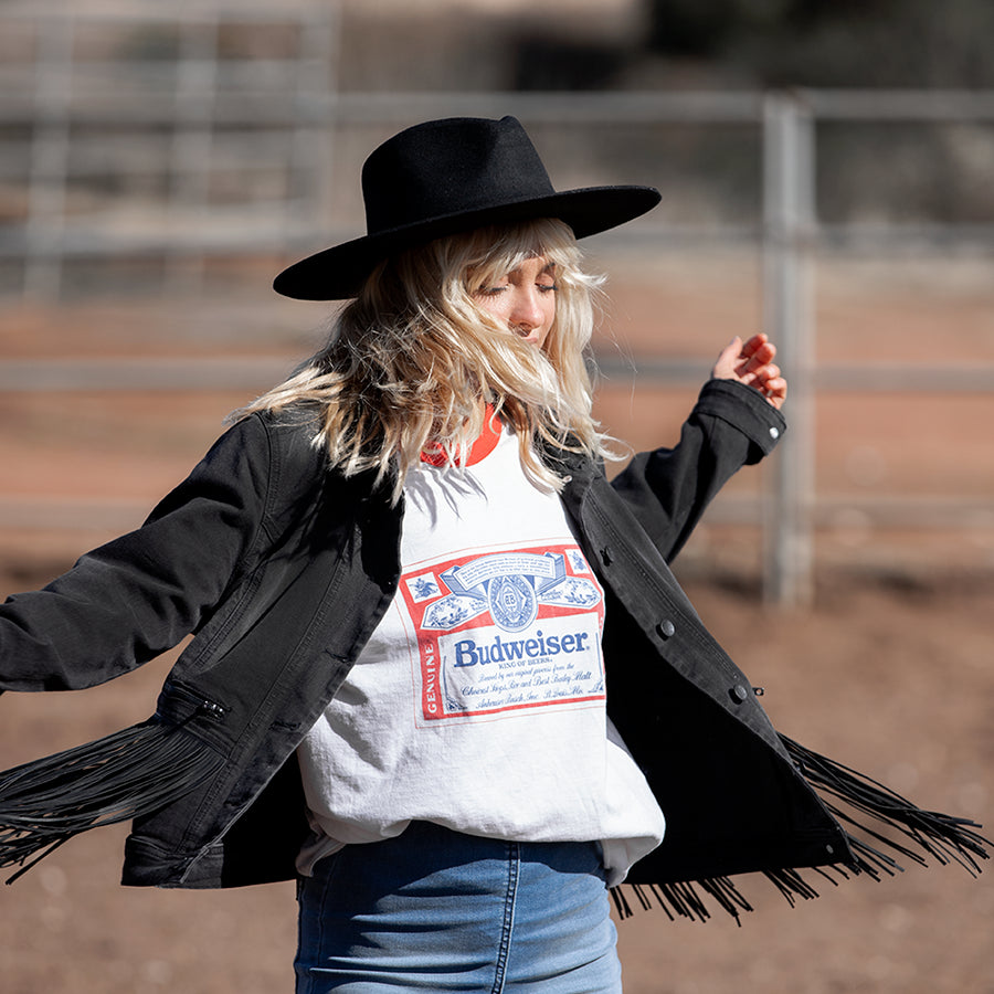 Person wearing a black coat with fringes, white t-shirt with Budweiser logo, and black hat outdoors.