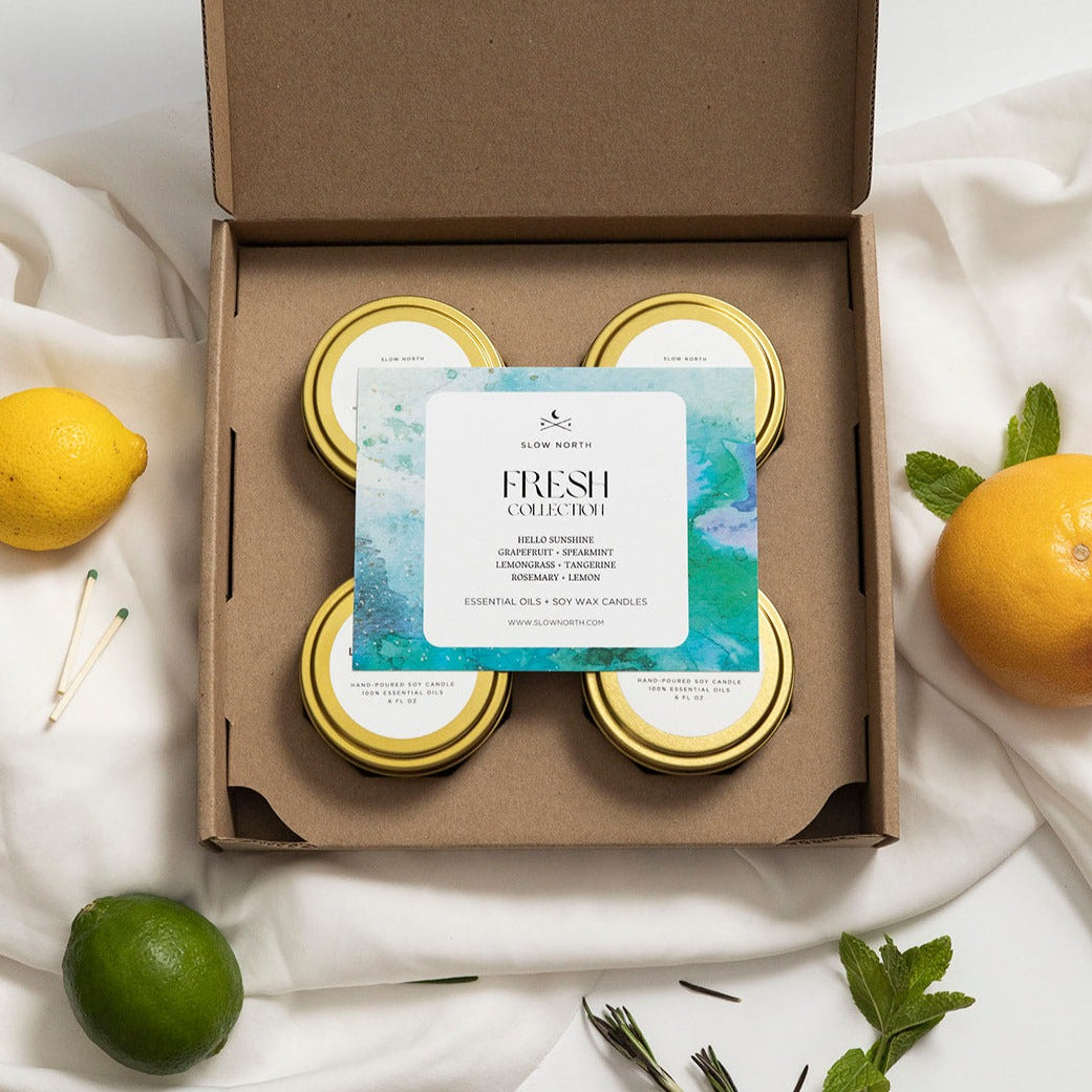 Box of candles with a card labeled 'Fresh Collection' on a white fabric background with fruits and herbs.
