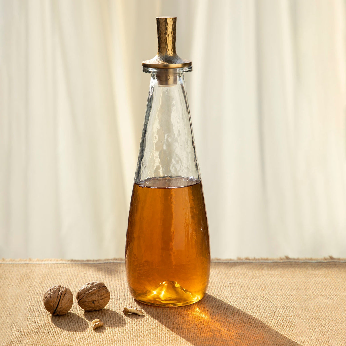 A clear glass decanter filled with amber-colored liquid, featuring a tall, hammered aluminum stopper with an antiqued brass finish, placed on a table with a curtain background.