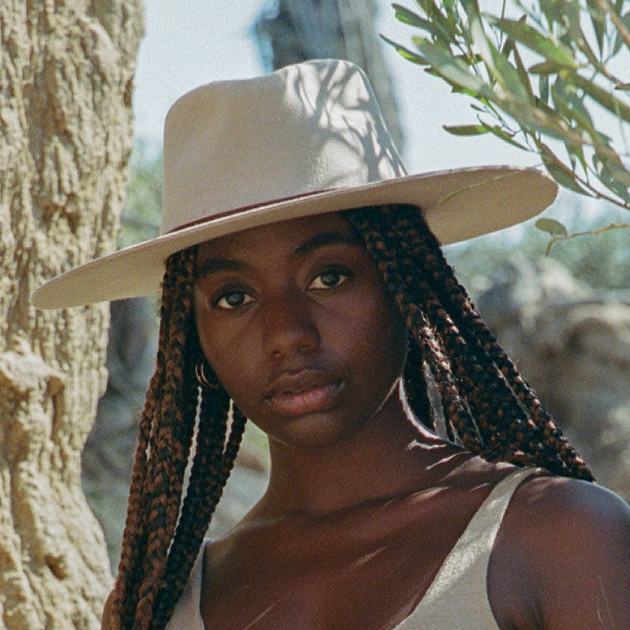 Woman wearing a beige hat with braided hair, standing outdoors with trees in the background