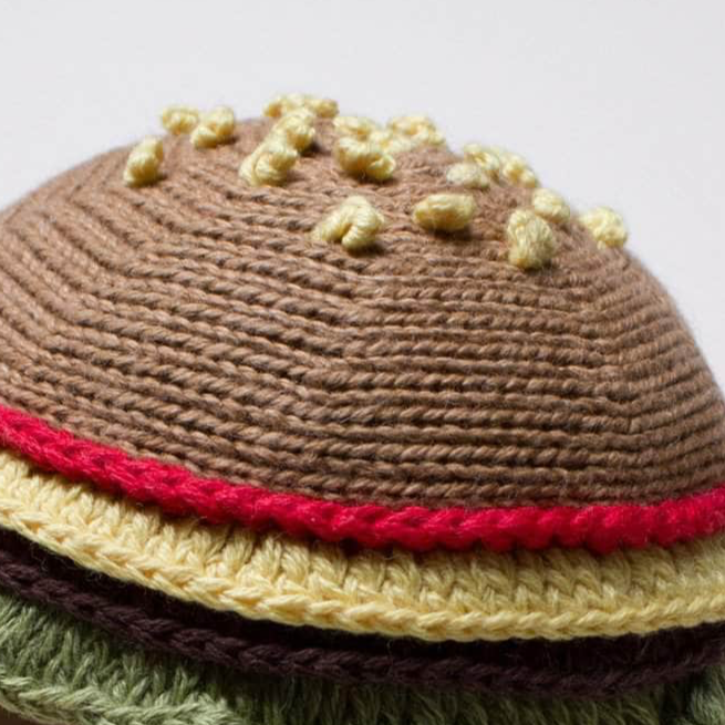 Knitted toy resembling a hamburger with brown bun, yellow cheese, red tomato, and green lettuce on a white background.