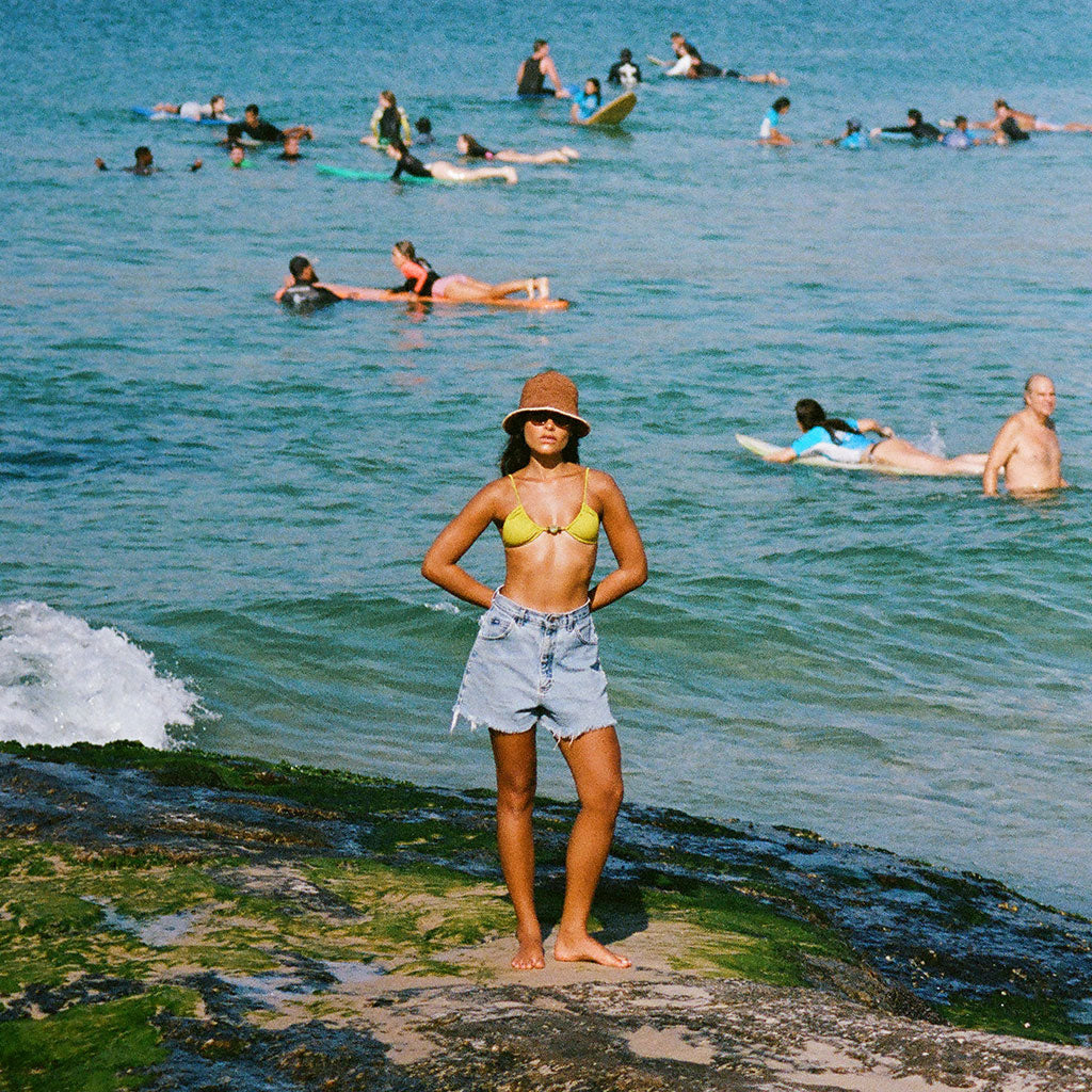 Woman in a yellow bikini top and denim shorts standing on a beach with people swimming and surfing in the ocean.