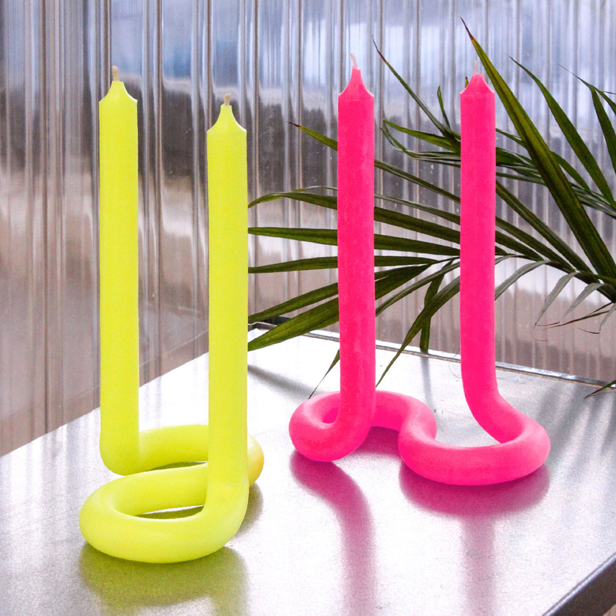 Two pairs of neon yellow and pink candles on a reflective surface with a blurred background.