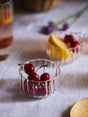 Clear glass bowls with cherries and fruit on a light fabric surface.
