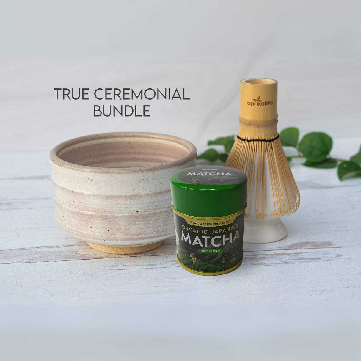 The Beige Ceramic Matcha Bowl, Bamboo Matcha Whisk, Whisk Holder, and Organic Japanese Matcha Set rest elegantly on a white wooden surface with leaves, highlighting the beauty of Japanese pottery.