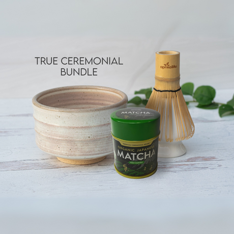 The Beige Ceramic Matcha Bowl, Bamboo Matcha Whisk, Whisk Holder, and Organic Japanese Matcha Set rest elegantly on a white wooden surface with leaves, highlighting the beauty of Japanese pottery.