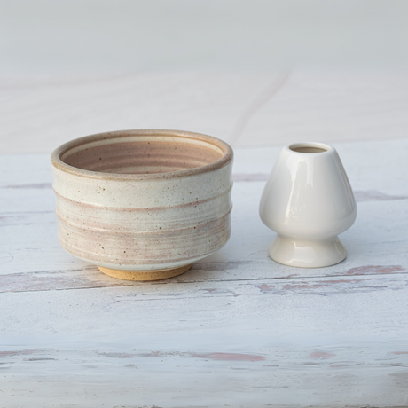 A Beige Ceramic Matcha Bowl from the Bamboo Matcha Whisk, Whisk Holder, and Organic Japanese Matcha Set rests gracefully alongside a small white vase on a light wooden surface.