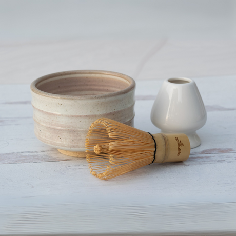 A Beige Ceramic Matcha Bowl, Bamboo Matcha Whisk, and Whisk Holder set on a white wooden surface exemplify elegant matcha accessories crafted from Minoyaki pottery.