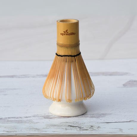 A bamboo matcha whisk on a white ceramic holder, part of the Beige Ceramic Matcha Bowl, Bamboo Matcha Whisk, Whisk Holder and Organic Japanese Matcha Set, crafted with Minoyaki pottery style, rests gracefully on a light wooden surface.