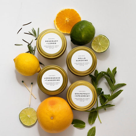 Four candles in gold tins with labels, surrounded by citrus fruits and herbs on a light background