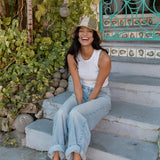 Woman sitting on steps outdoors, wearing a white top and light blue jeans, with greenery and a decorative door in the background.