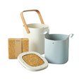 Set of kitchen utensils including a white container with wooden handle, gray pot, and cork items on a light surface.