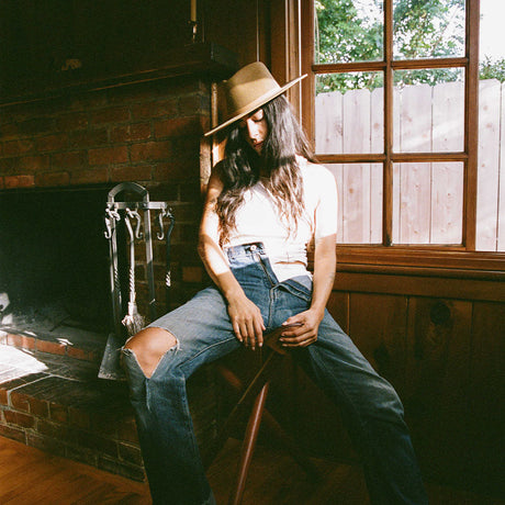Person wearing a hat and ripped jeans sitting by a window in a rustic room.