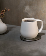 White mug with steaming coffee on a wooden surface next to a vase with flowers.