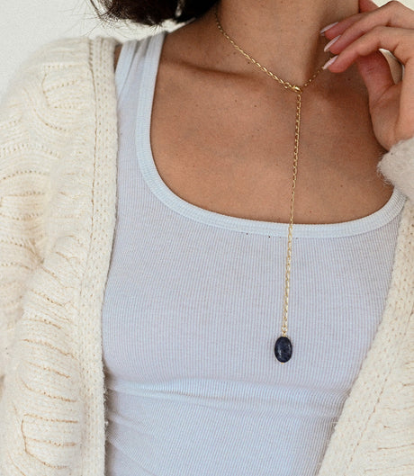 Person wearing a gold necklace with a dark pendant, dressed in a light blue top and beige cardigan.
