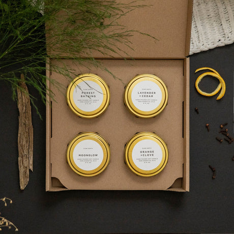 Four small candles in gold tins with labels in a cardboard box on a dark surface with herbs.